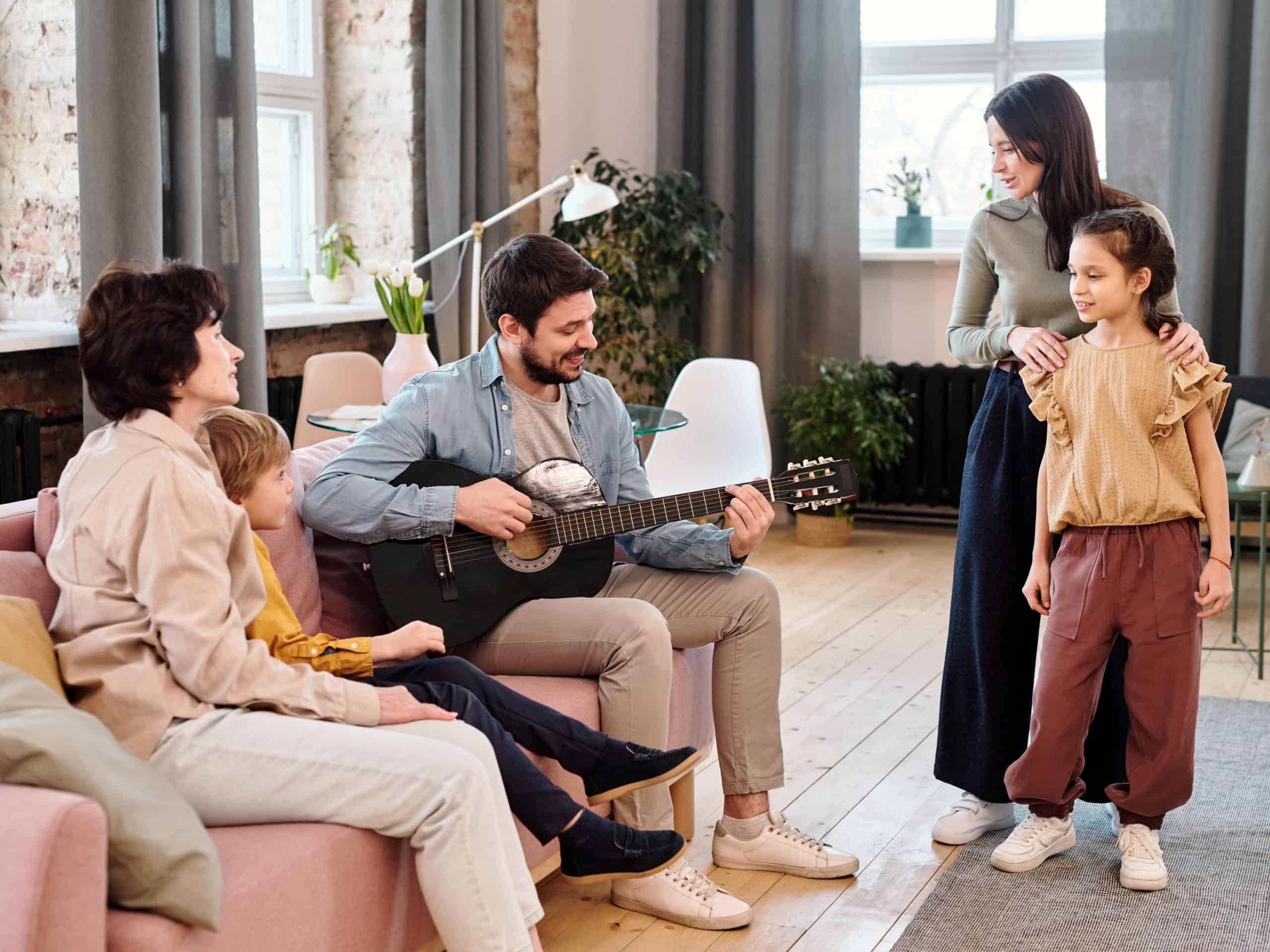 Happy young man playing guitar and singing while sitting on couch next to little boy and mature woman and wife and daughter in front